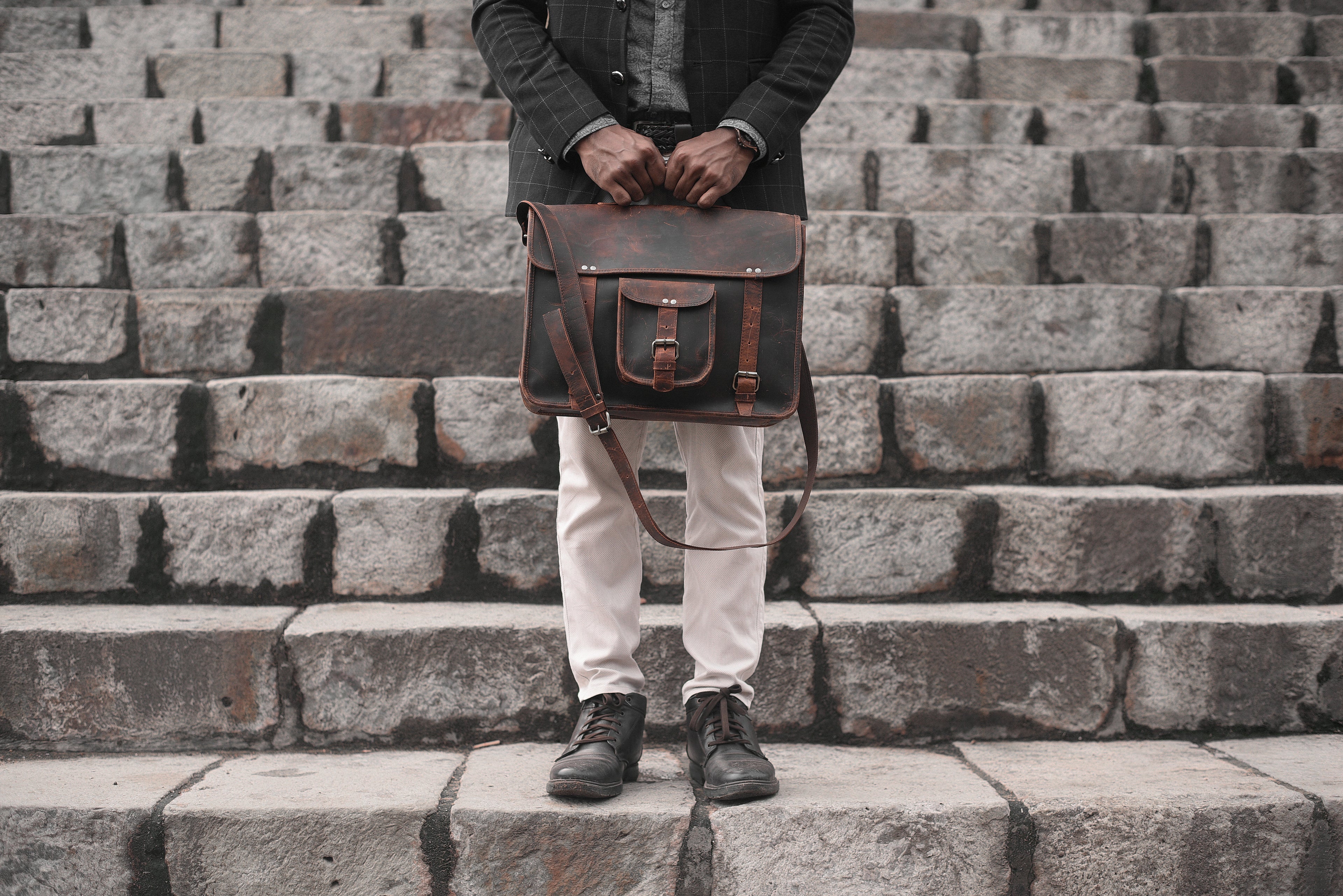 A man in a dark jacket and light pants stands on a stone staircase, holding a rustic, dark brown leather satchel with both hands.