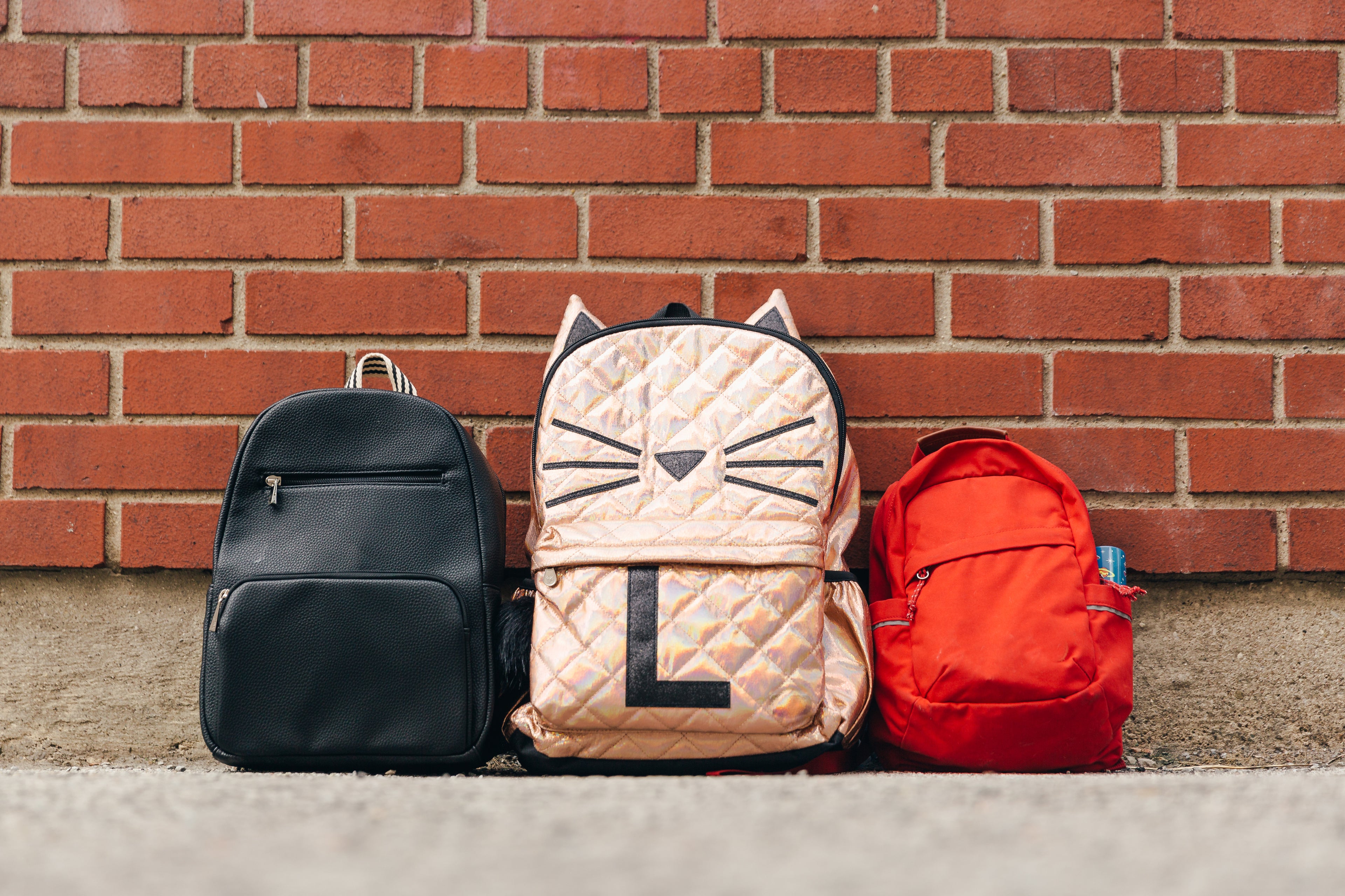 Three children's backpacks lined up on the ground against a brick wall. The backpacks are black, gold with a cat face design, and red.